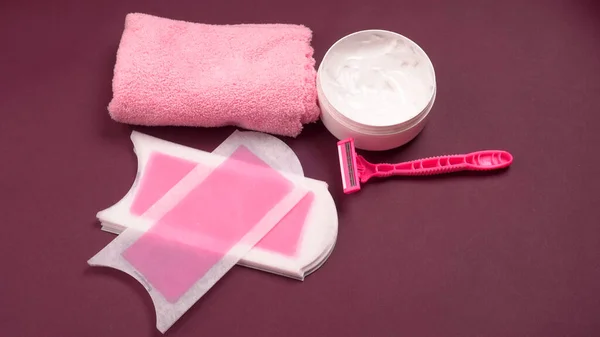 Closeup of wax strips with jar of cream and pink disposable razors for safe shaving of female skin on colorful towel, female hair removal concept