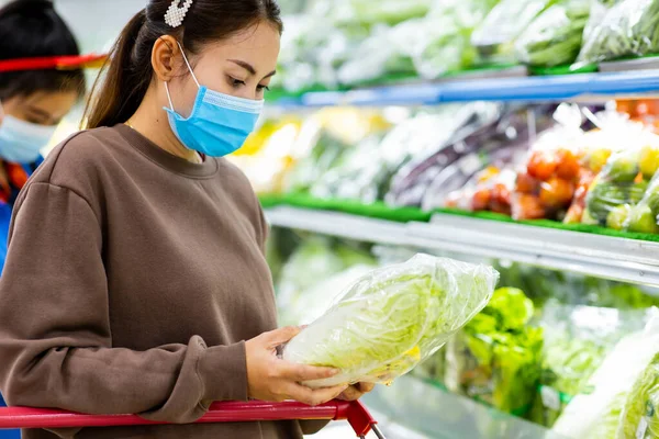 Young asian woman with protective mask pushing shopping cart for buying fresh vegetables in supermarket during virus COVID-19 outbreak. Concept for prevention COVID-19 Virus.