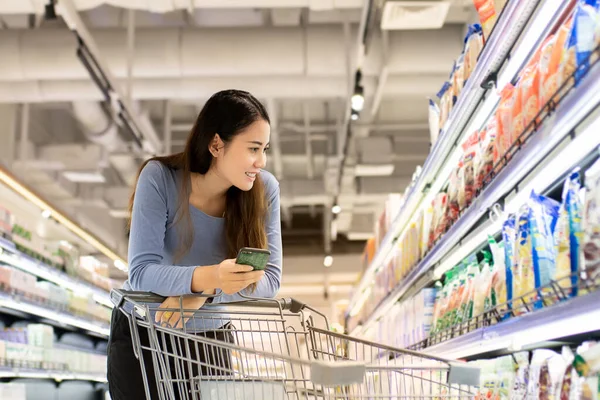 Güzel bir Asyalı kadın akıllı telefon kullanıyor ve market alışverişi için el arabasını itiyor. Alışveriş süpermarketi konsepti.