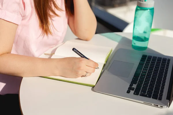 The hand of a girl in a pink blouse at the desk writes by hand in a notebook