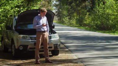 Arızalı arabanın yanında duran, akıllı telefon kullanan, hizmet bekleyen bir adam.