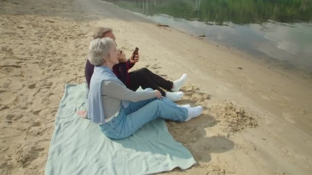 一对成熟的夫妇坐在沙滩上，通过智能手机听音乐 — 图库视频影像