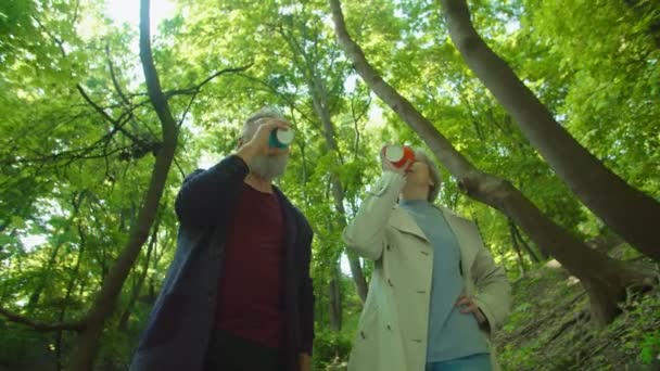 Atractiva pareja de ancianos con tazas de café caminando en el hermoso parque — Vídeos de Stock