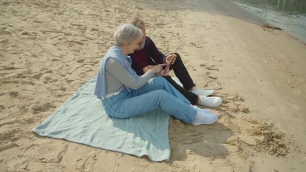 Ηλικιωμένο ζευγάρι που κάθεται στην άμμο, βλέποντας βίντεο ή φωτογραφίες μέσω tablet — Αρχείο Βίντεο
