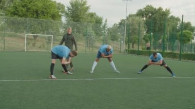 Koç açık hava takımına geniş bacaklı ön bükme egzersizi gösteriyor.