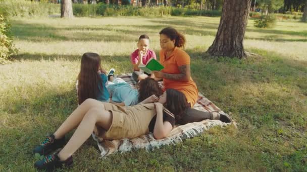Profesor de escuela leyendo libro a estudiantes de primaria de raza mixta al aire libre — Vídeos de Stock