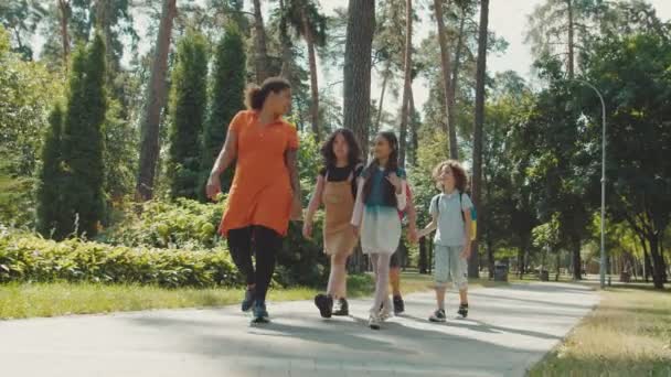 Jonge vrouwelijke leraar komt langs basisleerlingen in openbaar park — Stockvideo