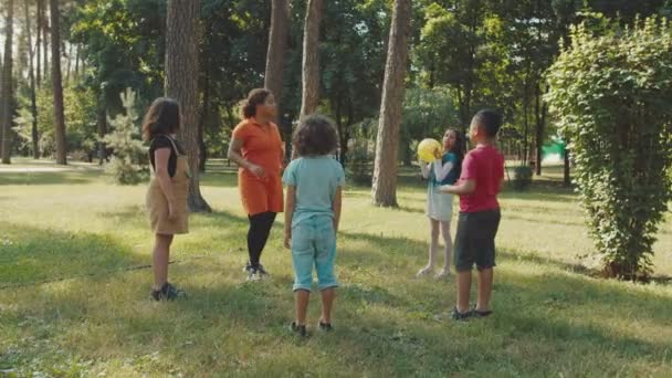 Los niños de la escuela jugando pelota con la maestra joven en el parque público — Vídeo de stock
