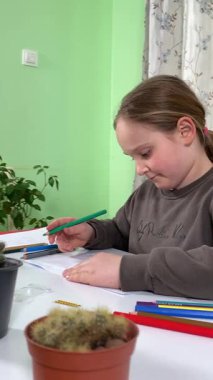 Genç kız evde yaratıcı aktiviteler yapıyor, hobisinin bir parçası olarak kalemlerle resim çiziyor. Odaklanmış ve ilham almış, sanatsal ifadenin günümüze getirdiği eğlence ve rahatlıktan zevk alıyor..