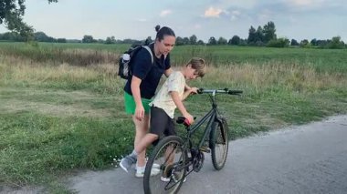 Bir annenin, küçük oğluna yeşil bir çayırda kırsal bir yolda bisiklet sürmeyi öğrettiği gibi doğada aile zamanı. Ebeveyn desteği, çocuk başarısı, neşe ve sıcak akşam güneşinde basit yaşam konsepti.