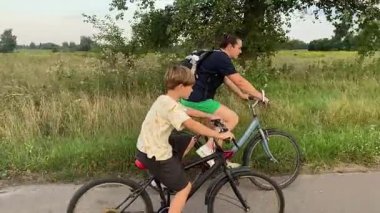 Anne ve oğlu gün batımında yeşil bir çayır boyunca bisiklet sürerken dışarıda mutlu bir aile zamanı. Sıcak akşam güneşi, doğa, rahatlama ve basit kırsal yaşam. Kaygısız ruh hali..