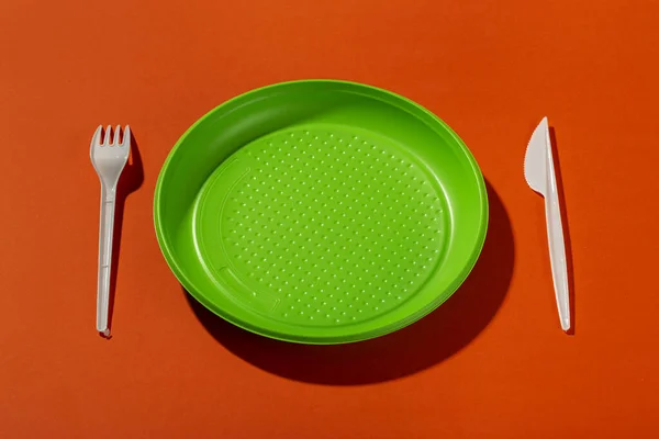 Plastic set of disposable tableware consisting of a plate, knife and fork on an orange background. Environmental concept. Ban single use plastic.Top view.