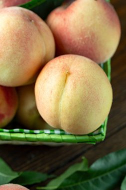 Tahta bir kırsal masada sepetin içinde taze şeftali hasadı. Şeftali yaprakları kompozisyonda mevcuttur. Organik meyve, vejetaryen yemeği..