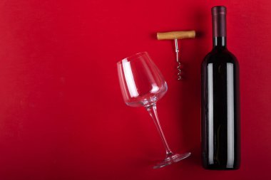 Bir şişe kırmızı şarap ve yanında tirbuşon olan boş bir şarap bardağı. Kırmızı arka planda kompozisyon. Romantik bir ruh hali. Mesaj için yer var. Üst görünüm.