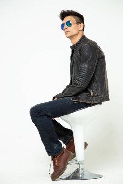 Beyaz arka planda gözlüklü şık bir genç stüdyoda bir sandalyede oturuyor.