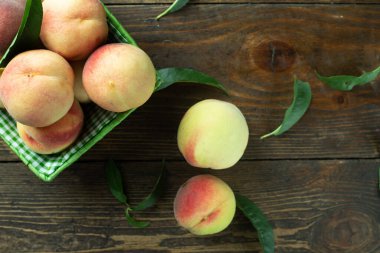 Tahta bir arka planda sepetin içinde olgun şeftaliler. Şeftali yaprakları masanın üstüne saçılmış. Meyve hasadı.