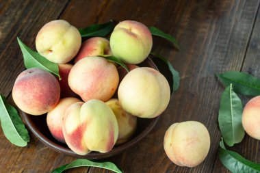 Ahşap bir kırsal masada, kil bir kasede taze şeftali hasadı. Şeftali yaprakları kompozisyonda mevcuttur. Organik meyve, vejetaryen yemeği..