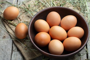 Ahşap arka planda kilden bir kasede tavuk yumurtası, saman etrafa saçılmış. Sağlıklı gıda konsepti. Çiftlik işçiliği ve bakımı.