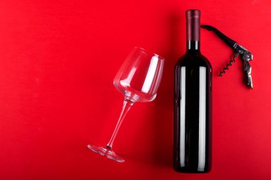 Bir şişe kırmızı şarap ve yanında tirbuşon olan boş bir şarap bardağı. Kırmızı arka planda kompozisyon. Romantik bir ruh hali. Mesaj için yer var. Üst görünüm.