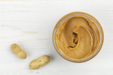 Beyaz ahşap bir masada fıstık ezmesi kavanozu ve yanında da fıstık. Mesaj için yer var. Üst görünüm.