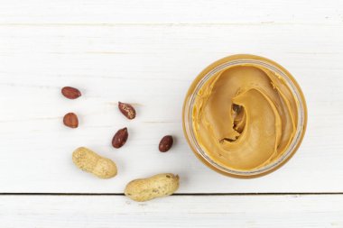 Fıstık ezmesi kavanozu beyaz ahşap bir masada, fındıklar etrafa saçılmış. Mesaj için yer var. Üst görünüm.
