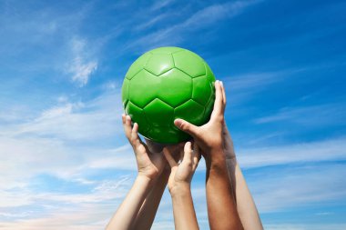 Yeşil bulutlu gökyüzünde kırmızı futbol topu tutan bir grup taraftar. Çocuklar ve yetişkinler el ele tutuşur.