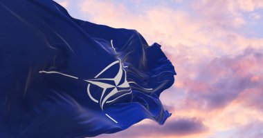 NATO bayrağı rüzgarda dalgalanırken bayrak direği gökyüzüne karşı güneşli bir günde bulutlarla.
