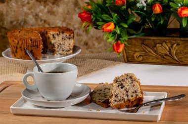 Buğday keki, çikolata ve kahveyle kahvaltı. Bir fincan kahve ve kekle kahvaltı masası. Arka plan pastası ve çiçekli sürahi..