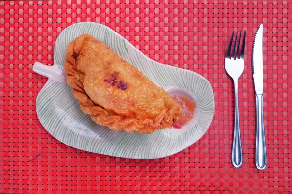 Volume de negócios frito chileno chamado Empanada, consistindo de pastelaria e recheio de atum, servido com um molho picante, tradicionalmente comido na Ilha de Páscoa, em uma tampa de mesa vermelha feita de plástico. — Fotografia de Stock