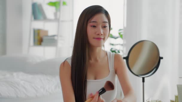 Egy gyönyörű ázsiai nő hurkolja a haját a tükör előtt. Egy pirosító ecsetet használ, hogy felerősítse az arcát..