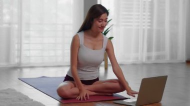Güzel bir Asyalı kadın yoga minderinde oturmuş internette yoga öğreten bir öğretmenle dizüstü bilgisayarına bakıyor. Meditasyon yapıyor ve yoganın başlangıç noktası olarak Lotus pozu veriyor..