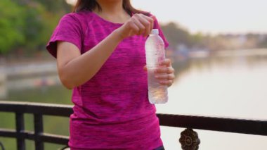 Asyalı kadınlar koşu yaptıktan sonra bir şişeden su içerek mola verirler. O, sabah parktaki gölün yanında koştu. Sağlık ve spor konsepti. Göl kenarına park et.