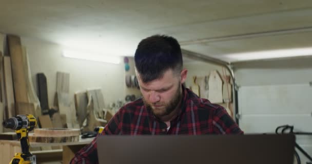 Man med skägg gör anteckningar framför öppen bärbar dator i snickeri verkstad — Stockvideo