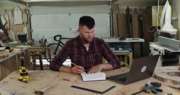 Man med skägg gör anteckningar framför öppen bärbar dator i snickeri verkstad — Stockvideo