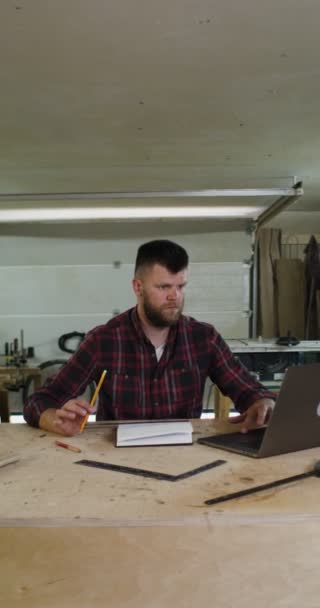 Uomo con barba prende appunti davanti al computer portatile aperto in falegnameria — Video Stock