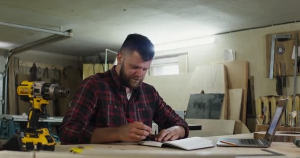 Un hombre europeo con barba haciendo notas en un cuaderno de taller de carpintería — Vídeos de Stock