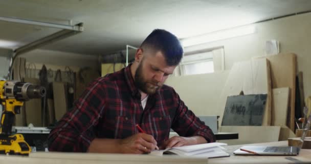 En europeisk man med skägg gör anteckningar i en anteckningsbok i snickeri verkstad — Stockvideo