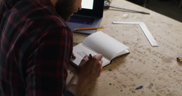 Un hombre europeo con barba haciendo notas en un cuaderno de taller de carpintería — Vídeos de Stock