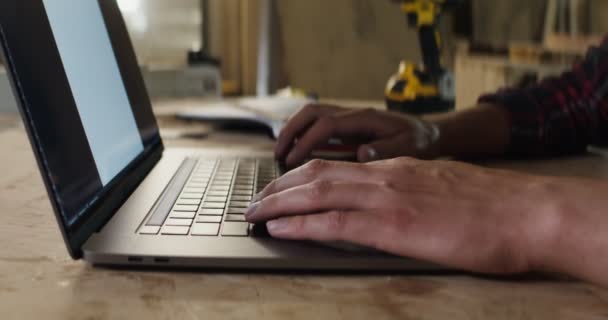 Man with fair skin is typing on laptop, hands close-up, in a carpentry workshop — Stock Video