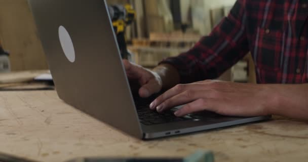 Homem com pele clara está digitando no laptop, mãos de perto, em uma oficina de carpintaria — Vídeo de Stock