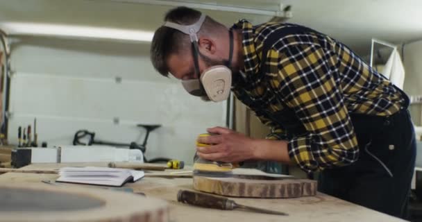 Man med europeiskt utseende i skyddande hörlurar och en mask bearbetar ett trä — Stockvideo