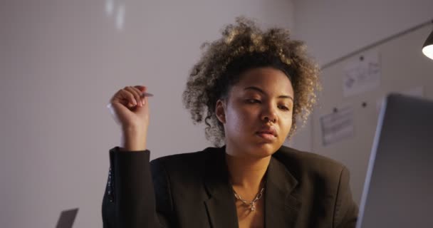 Chica de piel oscura en la chaqueta gira pluma, tipos y se ve en la pantalla en la oficina — Vídeo de stock