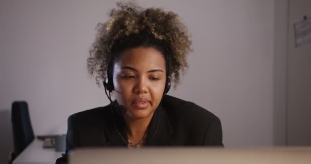 Chica africana en una chaqueta con auriculares con un micrófono habla y escucha — Vídeo de stock