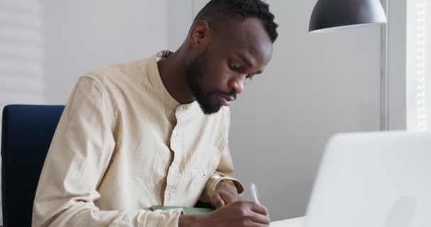 Negro hombre afro peinado hace notas en cuaderno, impresiones en el ordenador portátil en la oficina — Vídeo de stock