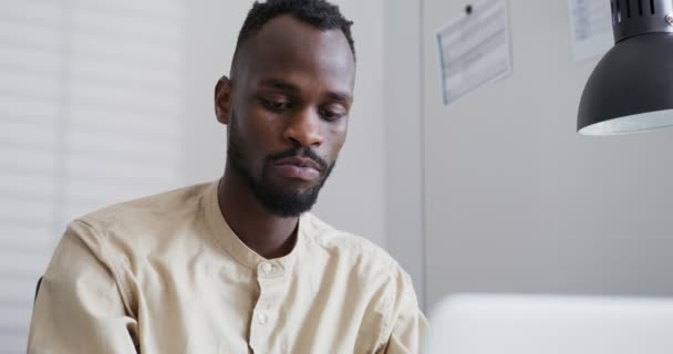 Zwarte man met een baard typen op een laptop, wrijft zijn kin in reflectie close-up — Stockvideo