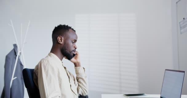 Svart kille med skägg talar i telefon medan du sitter på en öppen laptop på kontoret — Stockvideo