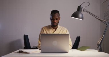 Ofisteki dizüstü bilgisayarda masa lambasının altında yazan siyahi bir adam.