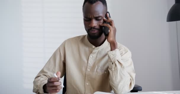 Noir gars avec une barbe parle sérieusement par téléphone tout en étant assis dans le bureau — Video
