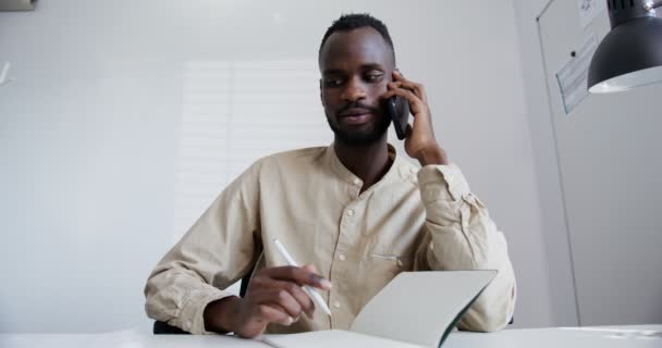 Svart kille med skägg talar allvarligt per telefon när han sitter på kontoret — Stockvideo