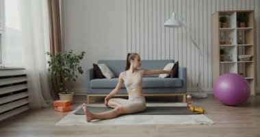 Spor kıyafetleri giymiş Avrupalı bir kız jimnastik minderinde oturarak egzersiz yapıyor.